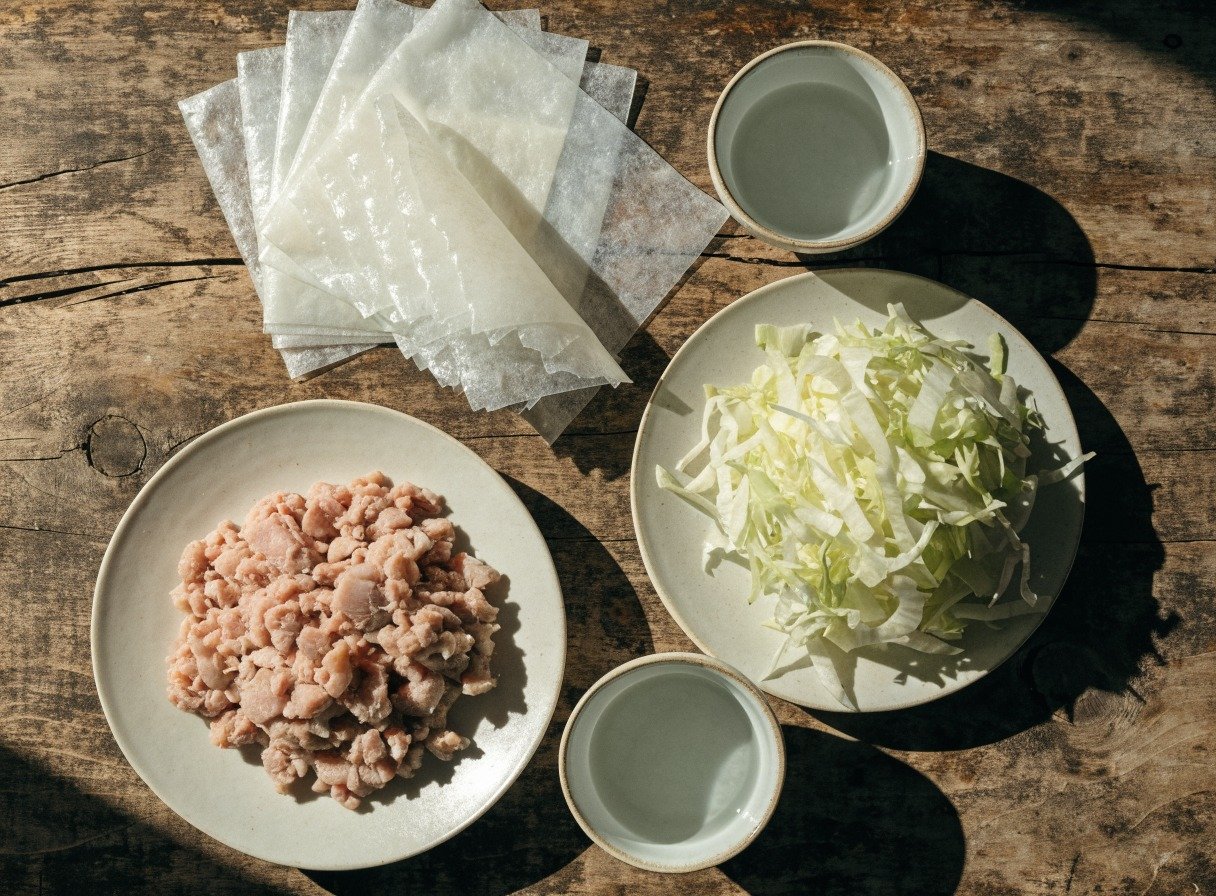 How do you keep rice paper from tearing when making dumplings? preparation close-up - Beautifully organized flatlay of rice paper wrappers, minced pork, chopped cabbage, and bowls of wat