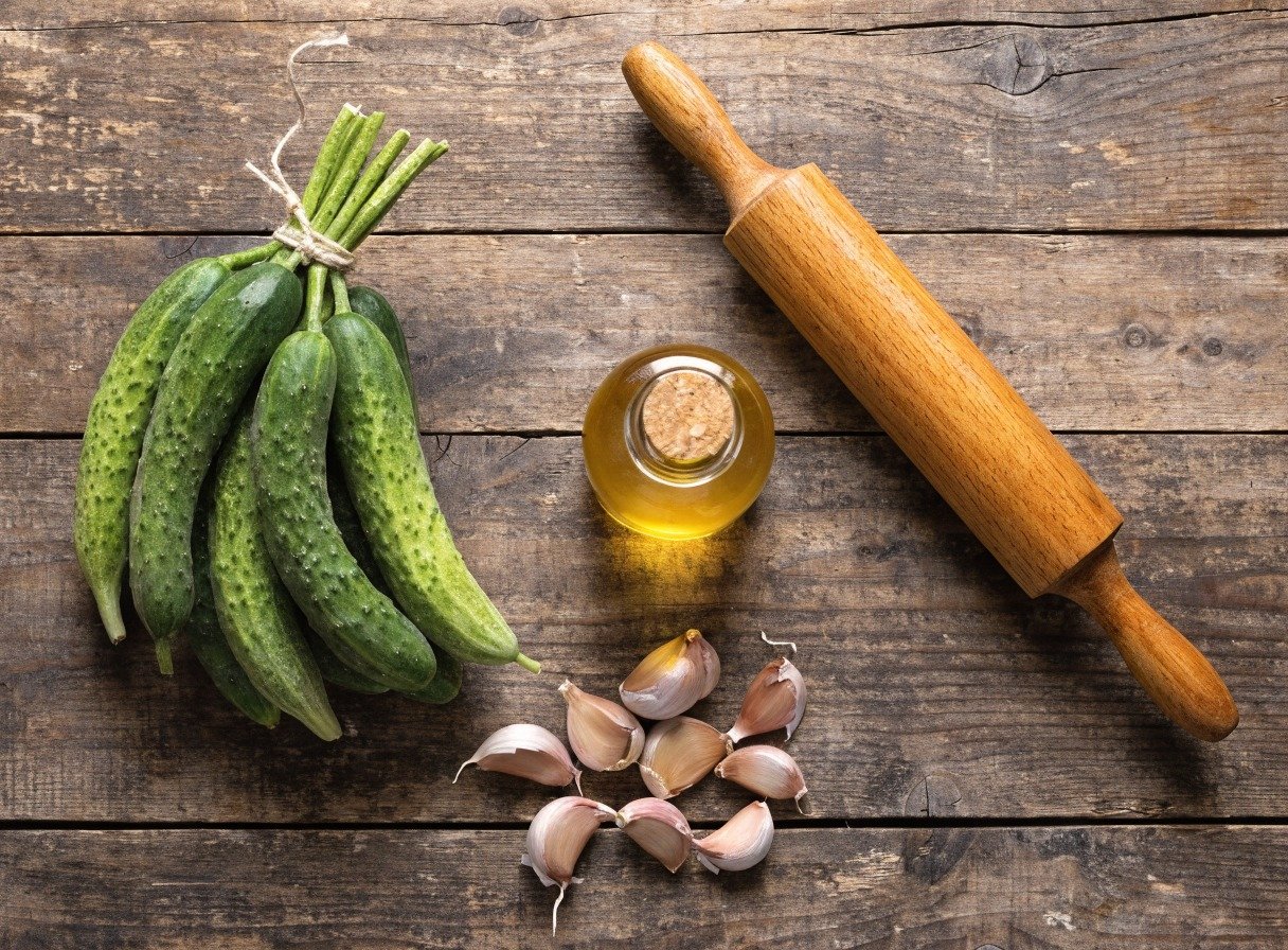 Why do you smash cucumbers in a salad? - Beautifully organized flatlay of fresh Persian cucumbers, garlic cloves, sesame oil, and a wooden rolling pin on a rusti