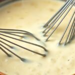 How do you fix a cream sauce that is too thick or grainy? recipe hero presentation - A close-up shot of a creamy white alfredo sauce in a copper saucepan, being whisked with a silver wh