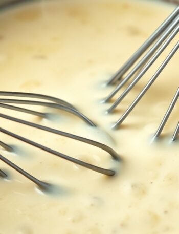 How do you fix a cream sauce that is too thick or grainy? recipe hero presentation - A close-up shot of a creamy white alfredo sauce in a copper saucepan, being whisked with a silver wh